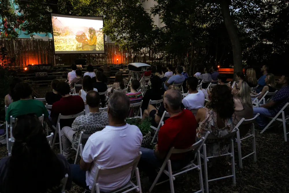 Publika na belim stolicama gleda film na platnu razapetom među drvećem, uveče u vrtu